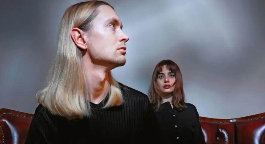 Łukasz Rozmysłowski z długimi blond włosami i kobieta z brązowymi włosami siedzą na skórzanej kanapie.