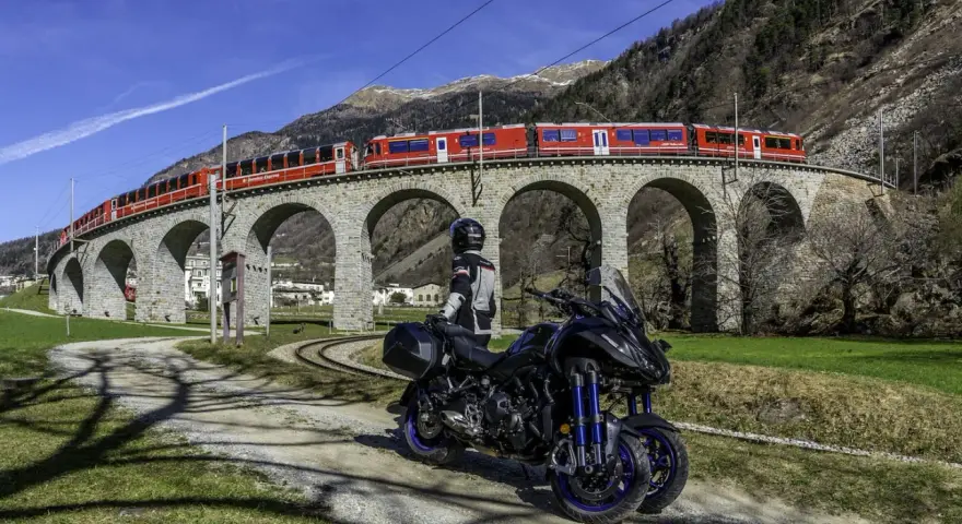 Motociclista e moto su strada sterrata, con un treno rosso che attraversa un viadotto ad archi in pietra. Paesaggio alpino con montagne sullo sfondo, vicino al passo della Novena.