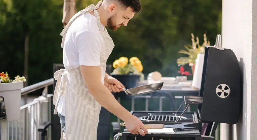 Mężczyzna w fartuchu przygotowuje grilla na balkonie, co pokazuje, że można robić grilla na balkonie.