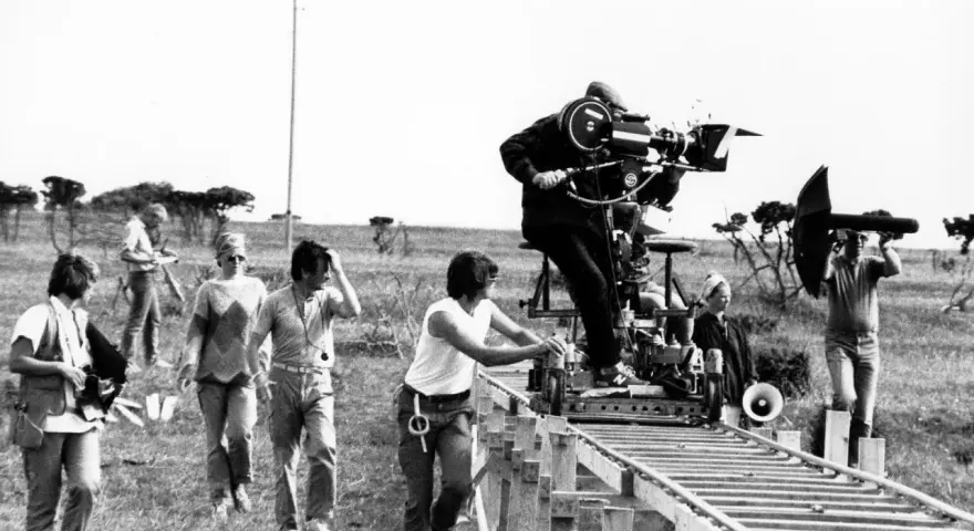 Sul set di un film, un operatore manovra una cinepresa su binari per un piano sequenza. Altri membri della troupe osservano e lavorano.