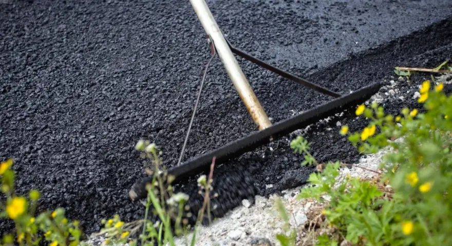 Verktyg sprider ny asfalt för att asfaltera uppfart. Grönska och gula blommor växer vid sidan.