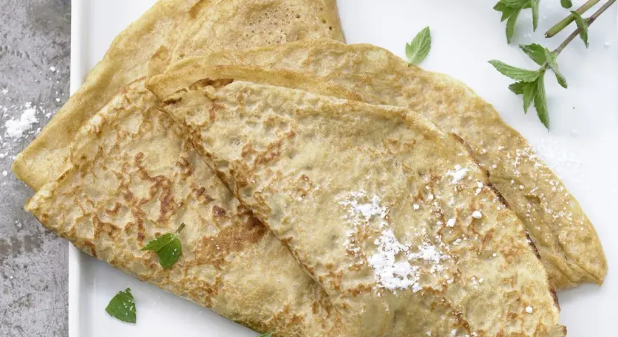 Drei goldbraune Dinkel Pfannkuchen, leicht mit Puderzucker bestäubt und mit frischer Minze garniert, auf einem weißen Teller.