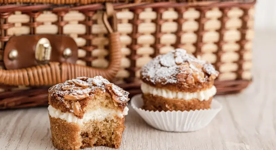 Zwei leckere bienenstich muffins mit Mandel-Topping und cremiger Füllung stehen vor einem Picknickkorb.