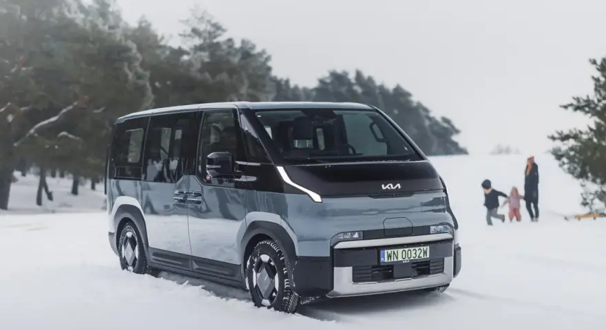 Szary elektryczny minivan Kia EV9 na śniegu, z rodziną w tle.