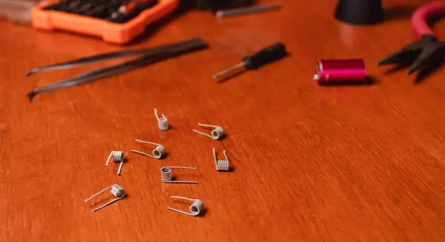 Varias resistencias comerciales de vapeo esparcidas sobre una mesa de madera, junto a herramientas como pinzas y un destornillador.