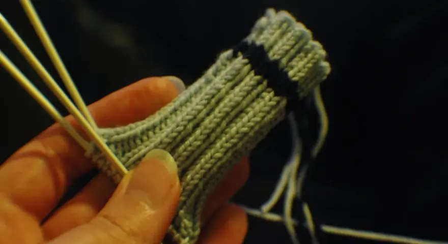 En hand håller stickor och ett ljusgrönt stickat tyg med en mörk rand. Det ser ut som att någon ska sticka avig maska.