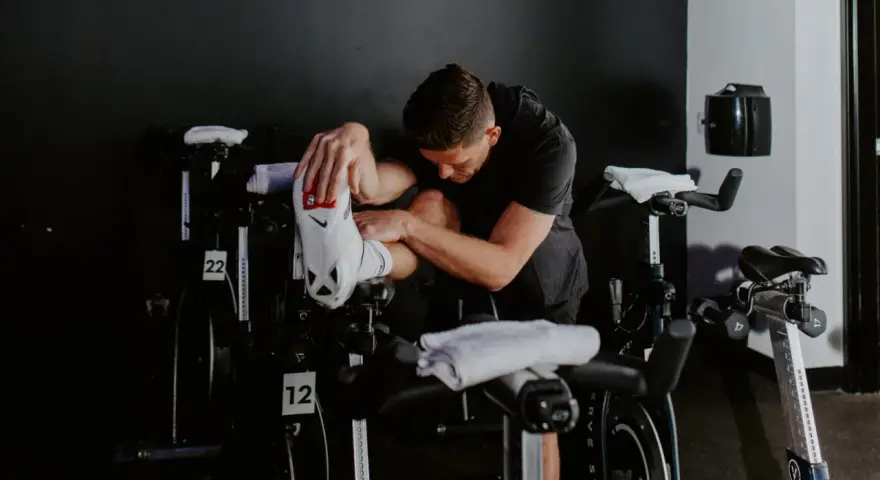 Man in black shirt stretches his outer hamstring by holding his foot, surrounded by stationary bikes in a gym.