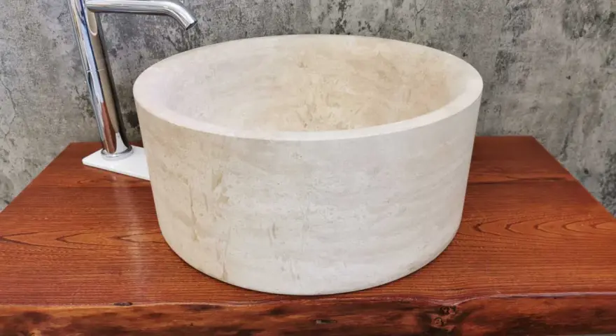 Lavabo cilindrico in pietra naturale chiara, con rubinetto cromato, su un piano in legno grezzo. Un esempio di come i nomi delle pietre naturali si trasformano in design unici.