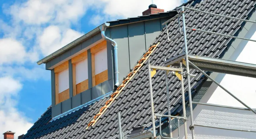 Bygga takkupa pågår. Ett hus med takpannor och byggställning, en ny takkupa med fönsteröppningar syns.