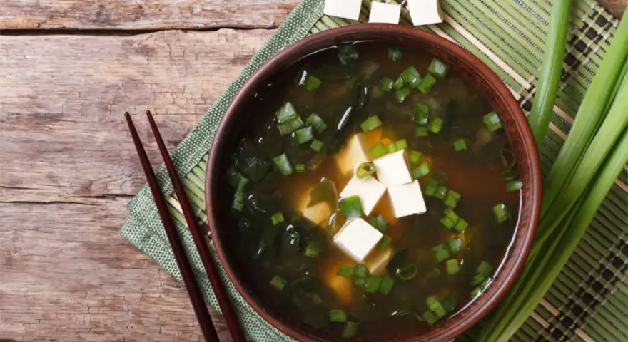 Aksamitne miso z tofu, wodorostami i szczypiorkiem, podane z pałeczkami na drewnianym blacie.