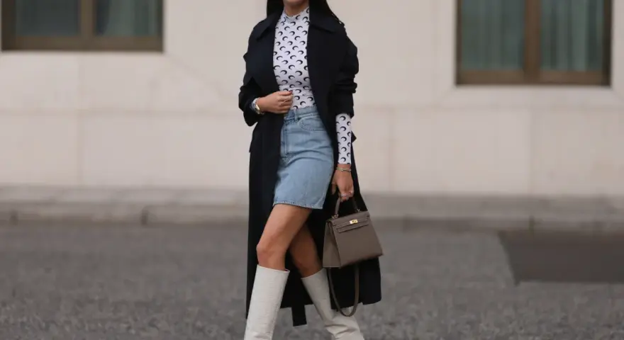 Outfit per uscire con un ragazzo: gonna di jeans, top a collo alto con stampa luna, trench nero, stivali bianchi alti e borsa grigia.