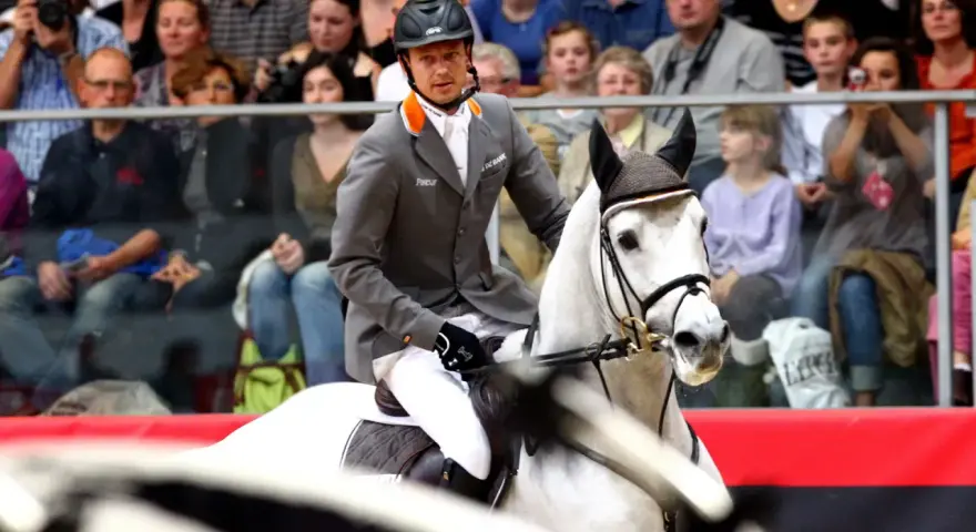 Un cavalier en tenue grise monte un cheval blanc, prêt pour la compétition. Le public est attentif.