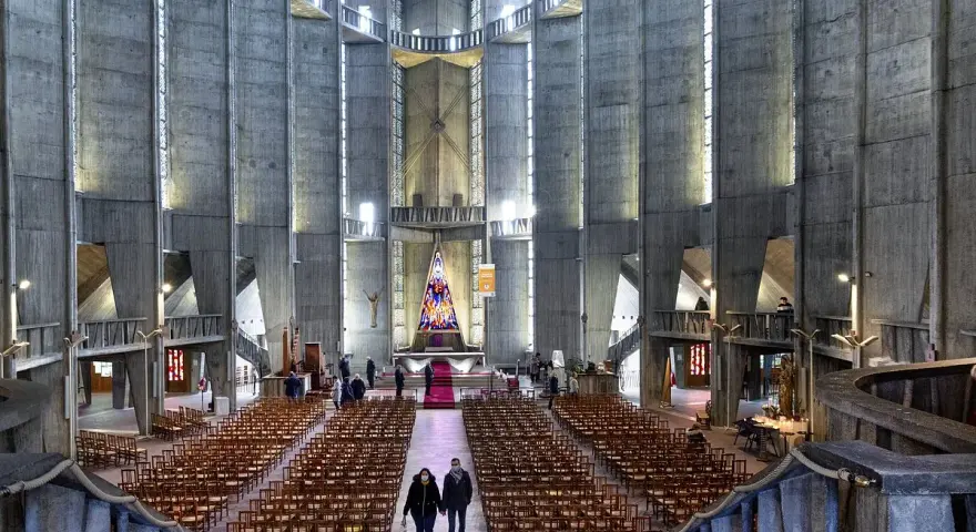 Intérieur de la cathédrale de Royan, avec ses murs en béton brut, ses rangées de chaises et son vitrail coloré au fond.