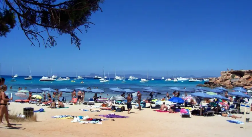 Playa con gente, sombrillas y barcos en el mar azul. Un día perfecto para ver qué ver en Formentera.