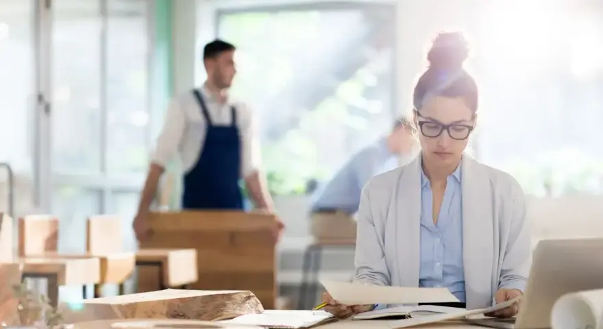 Kobieta w okularach analizuje dokumenty przy biurku, podczas gdy w tle mężczyzna w fartuchu pracuje przy drewnie. To jej staż z urzędu pracy.