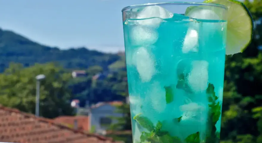 Orzeźwiający drink z blue curacao i sprite, z miętą i lodem, ozdobiony plasterkiem limonki, na tle zielonych wzgórz.
