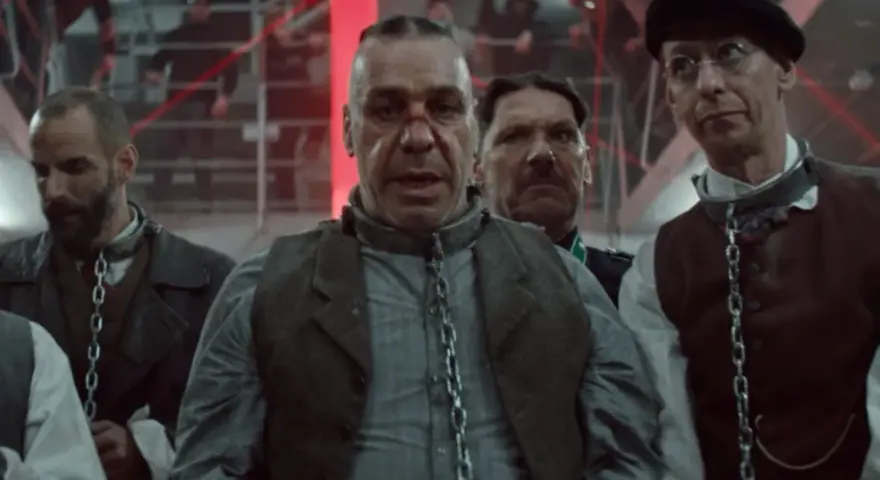 Rammstein - Deutschland: mężczyźni w łańcuchach, w tle czerwone światła i ludzie na schodach.
