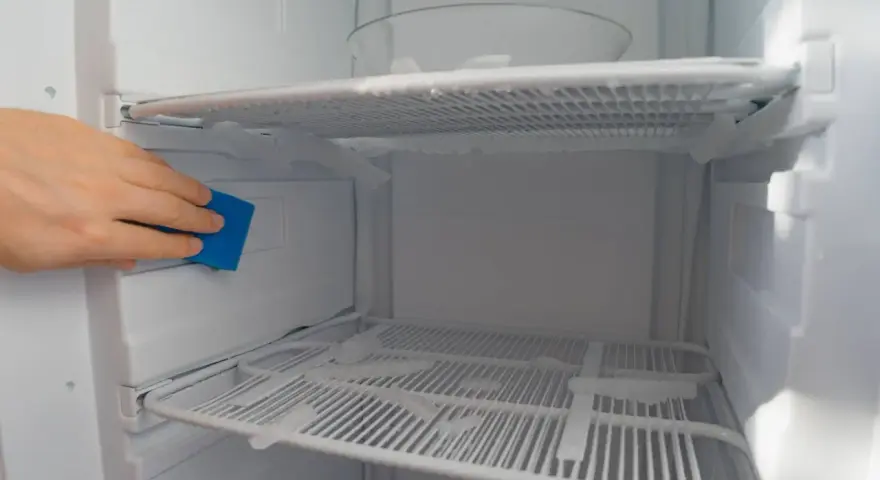 A hand uses a blue sponge to clean ice buildup inside a fridge freezer, showing a method for how to defrost fridge freezer without turning it off.