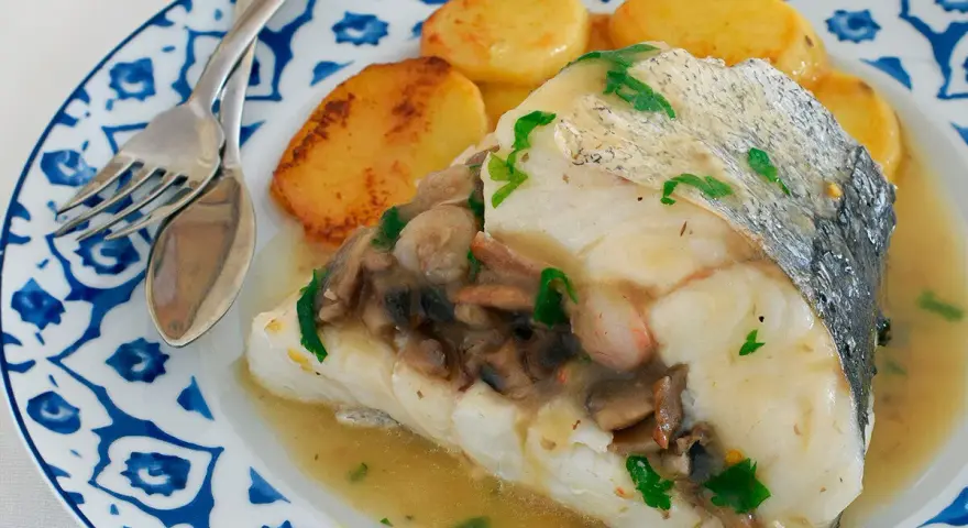 Deliciosa merluza con gambas y champiñones, servida con patatas doradas en un plato azul y blanco.