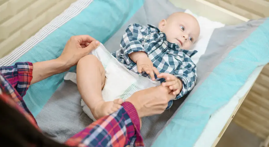Mama sprawdza, kiedy zmienić rozmiar pieluchy maluszkowi. Dziecko leży na przewijaku w kratkę.