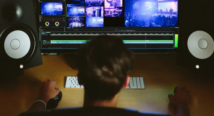 Osoba edytuje wideo na komputerze, otoczona przez jakie monitory studyjne. Widok zza pleców na ekran z wieloma kadrami i linią czasu.