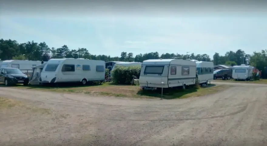 Ställplatser med husvagnar och bilar på Olles camping. Träd och blå himmel syns i bakgrunden.