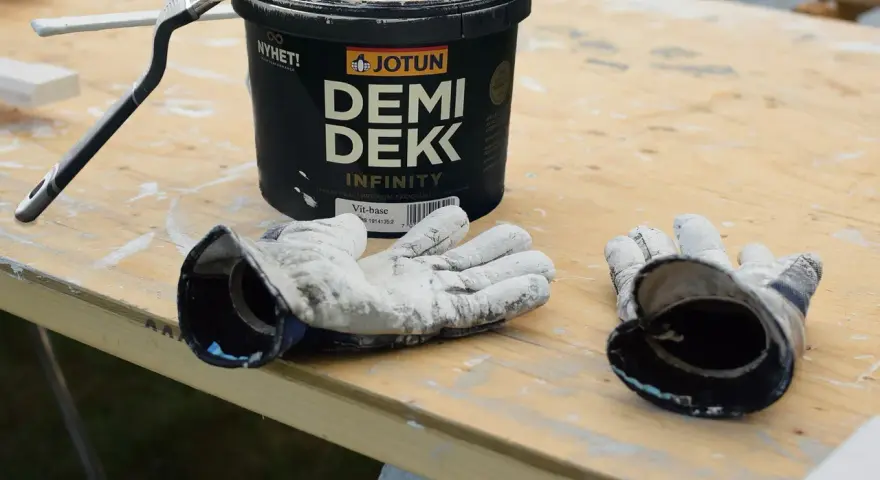 Förbereder för att måla utomhus på våren. En burk Jotun Demi Dekk, pensel och slitna handskar ligger på ett träbord.