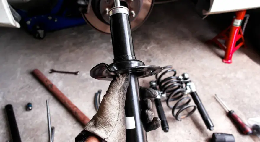 Mano guantata tiene un ammortizzatore nuovo, pronto per sostituire quelli scarichi dell'auto. Attrezzi e molle a vista.