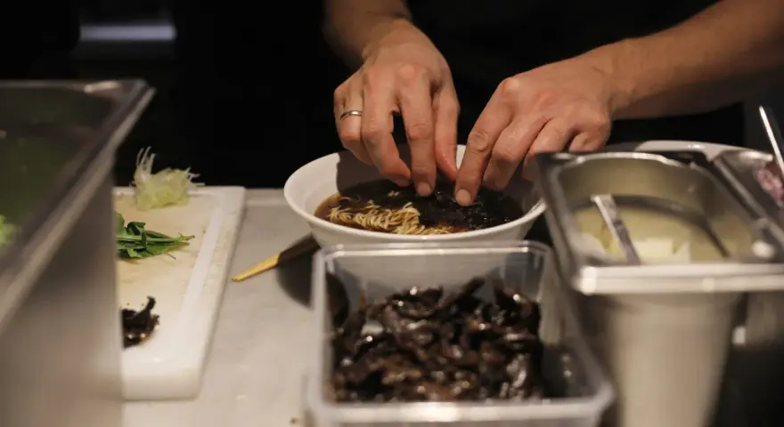 Hände garnieren eine Schüssel mit Ramen-Nudeln in Berlin. Daneben liegen Zutaten bereit.