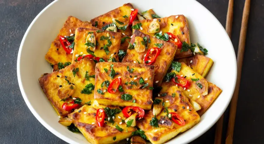 Chrupiące kawałki tofu satay, posypane chili, sezamem i ziołami, podane w białej misce z pałeczkami.