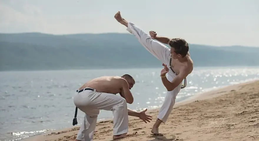 Dwóch mężczyzn ćwiczy capoeirę na plaży. Jeden wykonuje wysokie kopnięcie, drugi jest w pozycji obronnej. Widać ich płynny ginga styl gry.