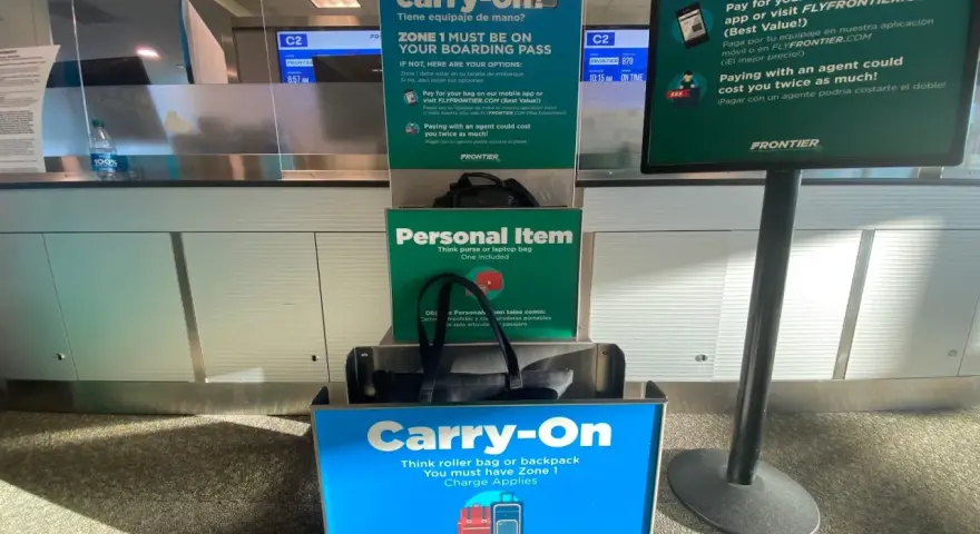 Frontier Airlines personal item size guide at the airport, showing a tote bag fitting in the "Personal Item" box and a roller bag in the "Carry-On" box.