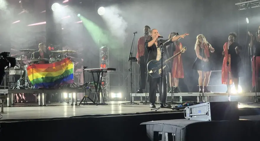 Wokalista z gitarą na scenie, obok chórki i perkusista z tęczową flagą. Koncert podczas festiwalu trzech kultur.