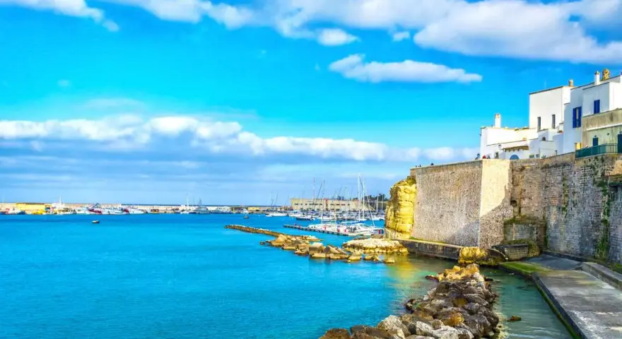 Un incantevole cammino del Salento: un porto con barche a vela, case bianche su scogliere dorate e un mare azzurro sotto un cielo limpido.