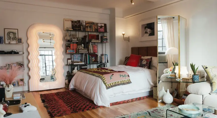 Ideas para decorar habitación estudio: cama blanca, espejo ondulado, estanterías con libros y arte, alfombras coloridas y un sofá acogedor.