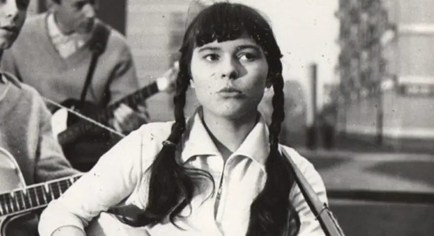 Młoda Karin Stanek z warkoczami śpiewa, grając na gitarze. W tle zespół muzyczny.