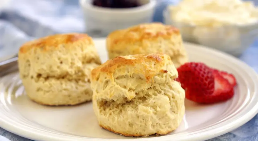 Tre gyllene engelska scones på en vit tallrik med en jordgubbe, sylt och grädde i bakgrunden.