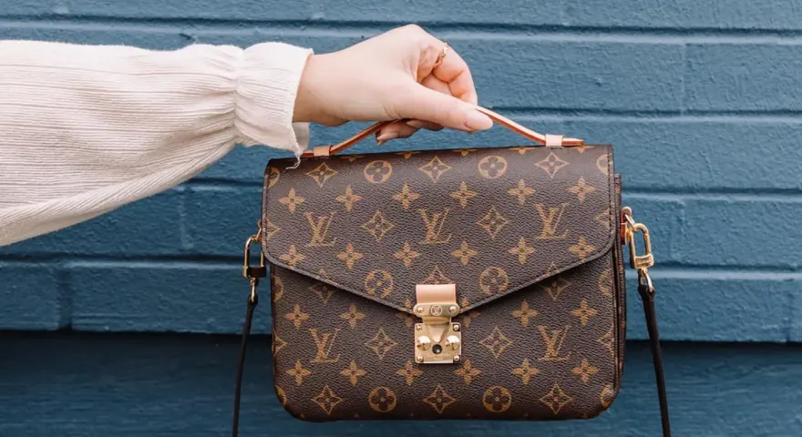 Una mano tiene una borsa Louis Vuitton con il classico monogramma. Un'icona di stile, che ci ricorda dove louis vuitton nasce: dalla maestria artigianale francese.