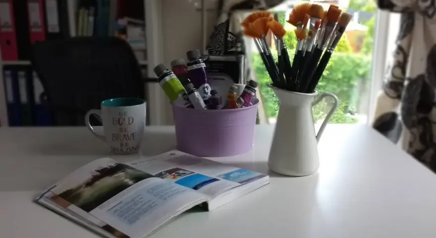 Pędzelki malarskie w dzbanku, tubki farb w fioletowym wiaderku, kubek z napisem i otwarta książka.