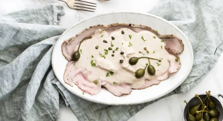 Eleganckie vitello tonnato z kremowym sosem, kaparami i natką pietruszki na białym talerzu, obok srebrnych sztućców i lnianej serwetki.