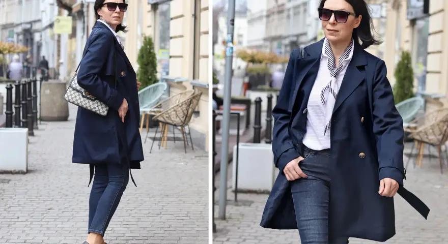 Elegancka kobieta w granatowym trenczu damskim, białej bluzce w paski i jeansach spaceruje po brukowanej ulicy.