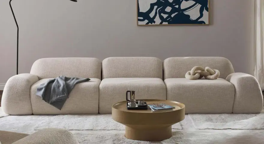 Divano modulare in tessuto bouclé color crema, con cuscino decorativo a forma di catena. Il materiale del divano è morbido e invitante, perfetto per un salotto moderno.