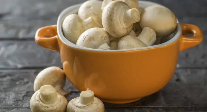 Pomarańczowa miseczka pełna świeżych pieczarek, jakby w ciąży z nowymi smakami. Kilka pieczarek leży obok na drewnianym blacie.