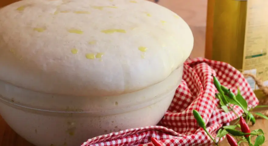 Impasto lievitato per pizza con lievito di birra, soffice e profumato, pronto per essere infornato. Olio e peperoncini completano la scena.