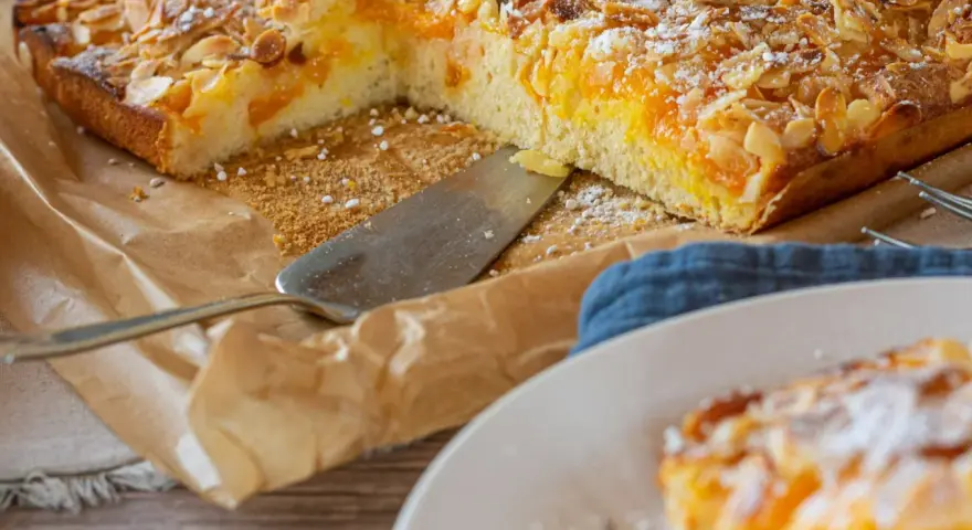 Duże ciasto z brzoskwiniami i migdałami, posypane cukrem pudrem, na papierze do pieczenia.