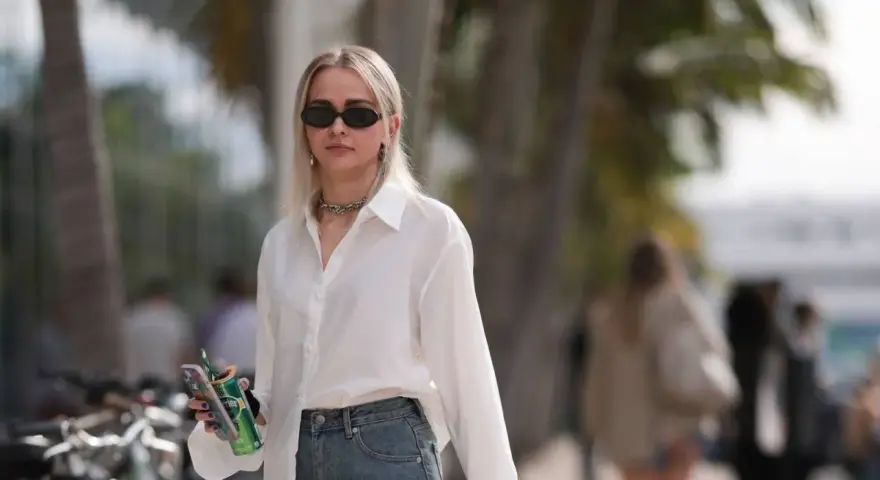 Donna con occhiali da sole, collana, camicia bianca e jeans, tiene in mano una lattina verde e un telefono.