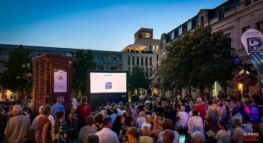 Ein lauer Sommerabend in Berlin: Menschen genießen ein Open-Air-Kino-Erlebnis vor einer Leinwand mit dem Logo "Lux Augenoptik".