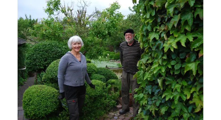 A smiling woman and a man stand in a lush garden with manicured bushes and trees.