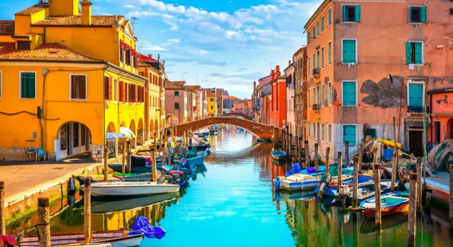 Colorate case affacciate su un canale con barche ormeggiate. Un ponte ad arco collega le due sponde. Un'immagine che ispira su cosa fare a Chioggia.