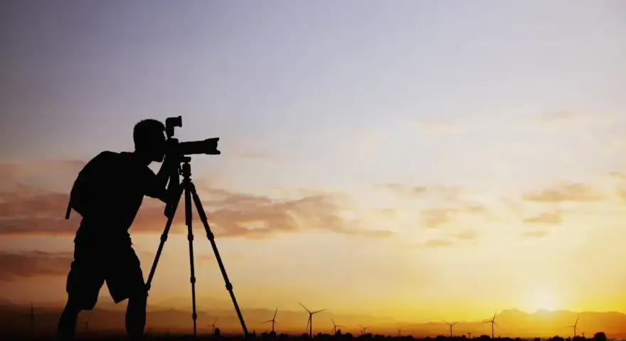 Sylwetka fotografa z aparatem na statywie na tle zachodzącego słońca i farmy wiatrowej. Zastanawiam się, ile bierze fotograf za 18 takich ujęć.
